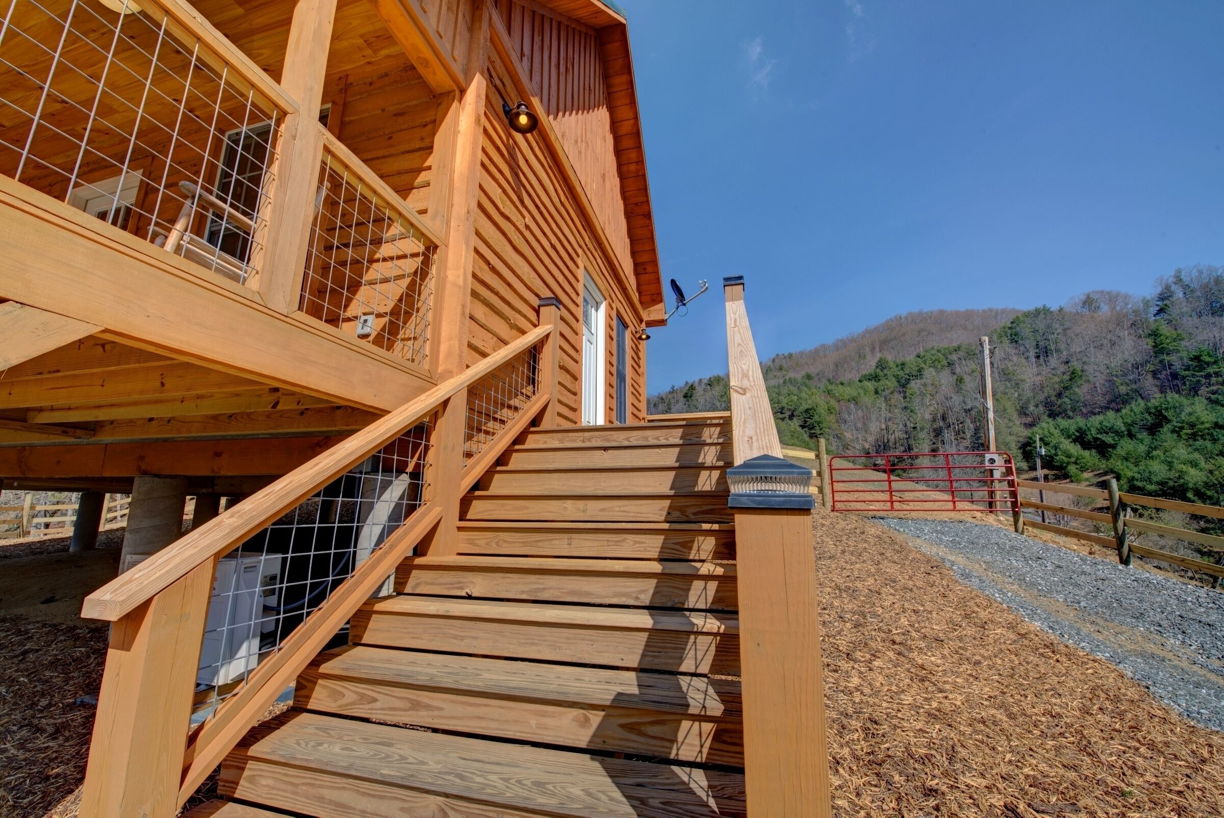 Un escalier en bois mène à un bâtiment doté d’une rampe métallique, et en arrière-plan, on aperçoit une colline boisée.