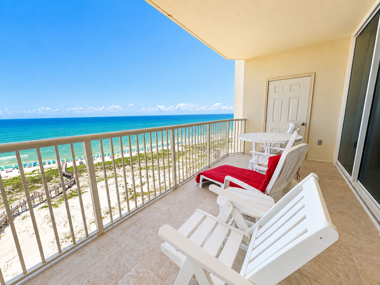 A balcony with a view of the beach and ocean, white furniture, and a door.