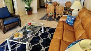Ein Wohnzimmer mit braunem Sofa, blauem Sessel, Glastisch und einem Essbereich mit Korbstühlen. Im Hintergrund ist eine Küche zu sehen.