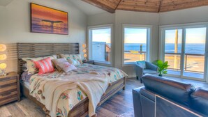 Une chambre à coucher comprenant un lit, des tables de chevet, une chaise et de grandes fenêtres donnant sur la plage.