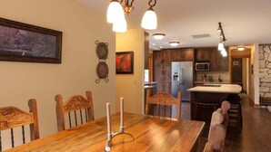 Un comedor con mesa de madera, sillas y lámparas colgantes. Se observa una cocina con armarios de madera y una pared de acento de piedra al fondo.
