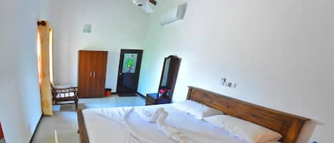 A hotel room with a large bed, wooden headboard, a ceiling fan, an air conditioning unit, a wooden wardrobe, and a small table with a chair.