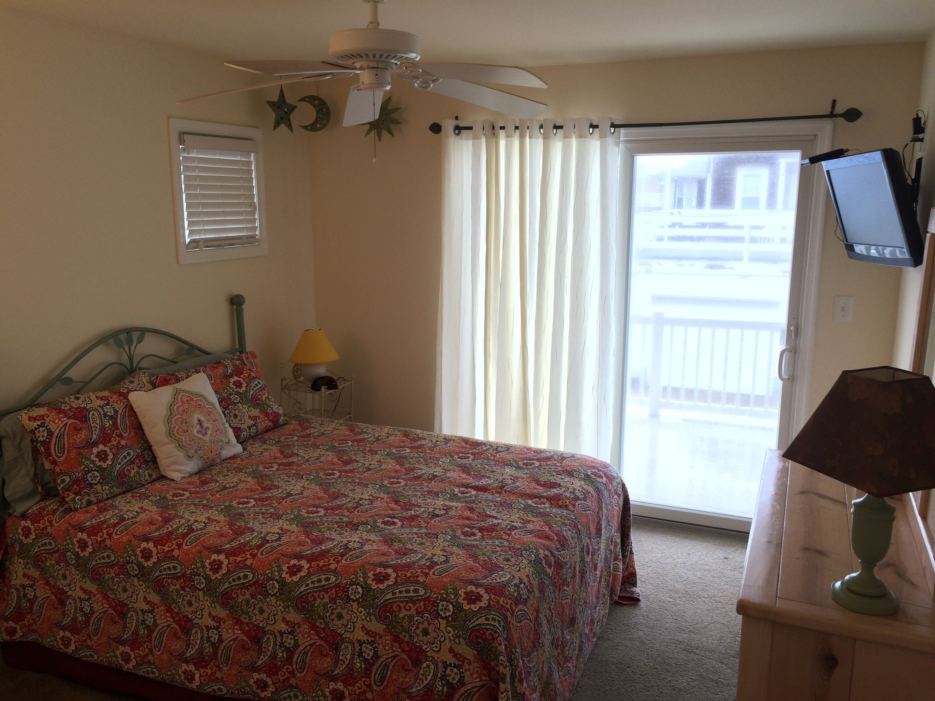 Une chambre à coucher avec un lit, un ventilateur de plafond, une table de chevet avec une lampe, un téléviseur posé sur un meuble et une fenêtre avec des rideaux.