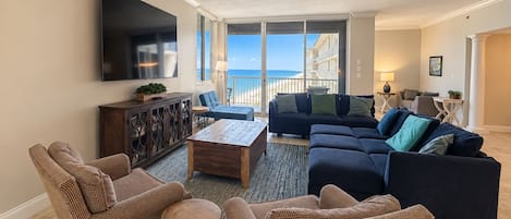 A living room with a large TV, a sofa set, a coffee table, and a view of the ocean.