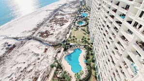 Ein Hochhaus-Hotel mit mehreren Balkonen, einem Poolbereich mit kleinem Pool und Whirlpool sowie einem Strandabschnitt mit einem Gehweg.