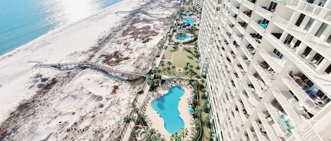 Un hotel de gran altura con múltiples balcones, un área de piscina con piscina pequeña y jacuzzi, y una zona de playa con un sendero para caminar.