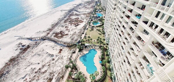 A high-rise hotel with multiple balconies, a pool area with a small pool and a hot tub, and a beach area with a walking path.