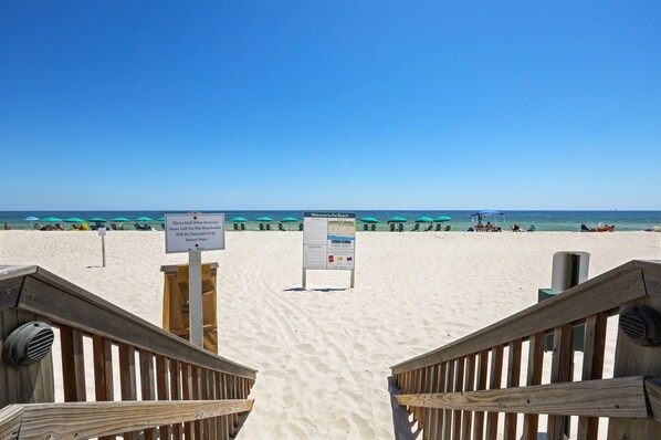 A beach entrance with wooden railings, a signboard, and beach umbrellas in the background.