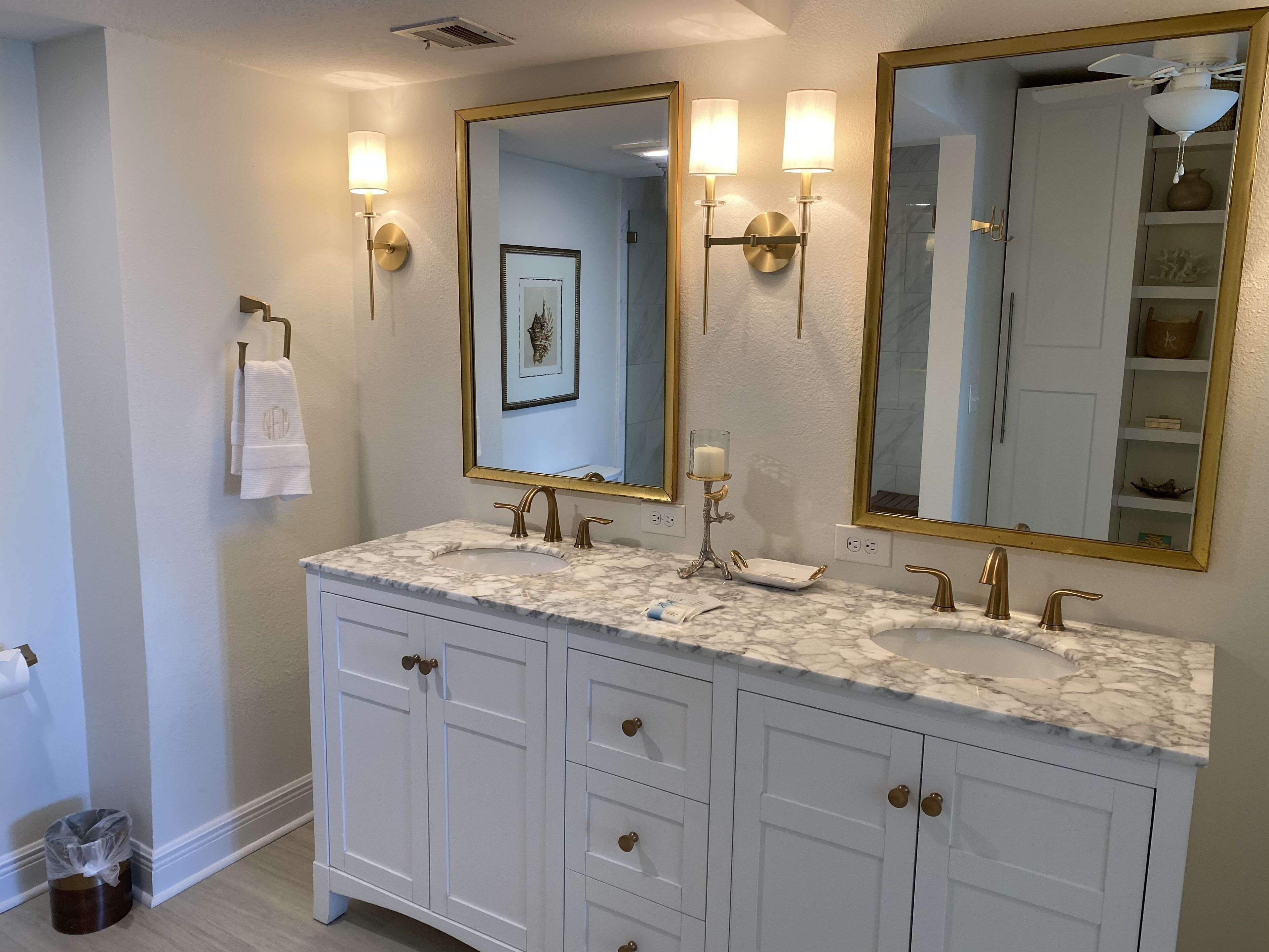 Un baño con dos lavamanos, muebles blancos, detalles dorados y espejos montados en la pared con apliques.