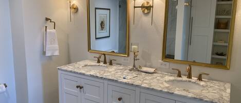 Un baño con dos lavamanos, muebles blancos, detalles dorados y espejos montados en la pared con apliques.