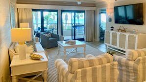 A hotel room with a seating area featuring striped sofas, a white side table with a lamp, a television mounted on the wall, and a balcony with a view of palm trees and the ocean.