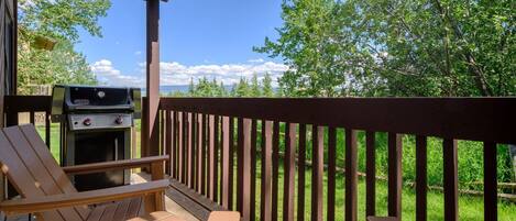 Une terrasse en bois avec un barbecue, deux fauteuils Adirondack et une vue sur les arbres et un ciel dégagé.