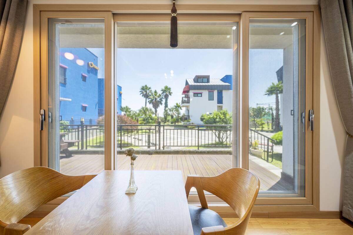 A room with a wooden table and chairs, a vase with a flower, and a view of a blue building and palm trees through a sliding glass door.