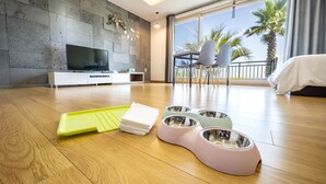 A modern living room with a flat-screen TV, pet bowls, and a dining area with chairs. - Jeju Blue & Sea (Seogwipo)