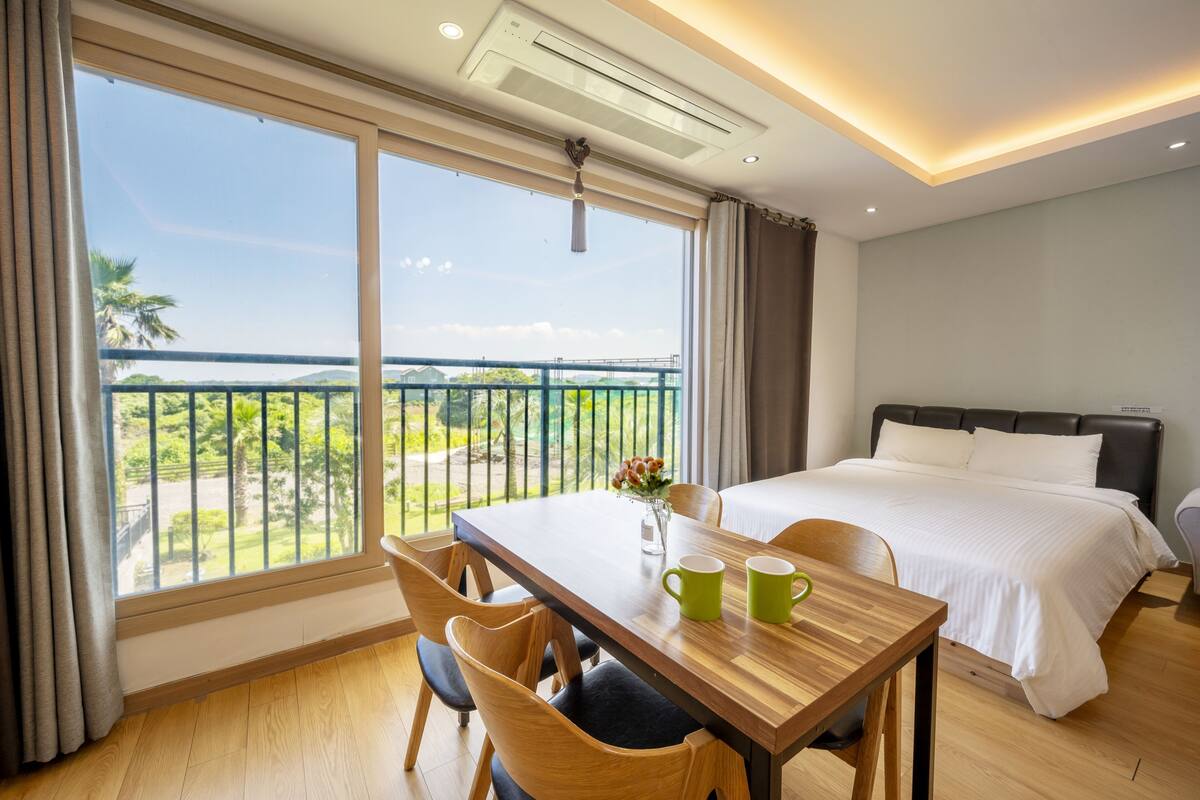 A hotel room with a balcony, a bed, a dining table with chairs, and a vase with flowers.