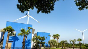 A hotel with a blue facade, palm trees, and wind turbines in the background. - Jeju Blue & Sea (Seogwipo)