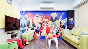 Ein Wohnzimmer mit einer großen Wandmalerei im Anime-Stil, einem Flachbildfernseher, einem gelben Sofa und Kinderspielzeug.