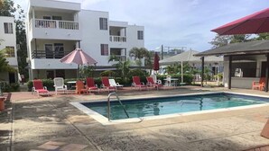 Une piscine entourée de chaises longues rouges et blanches, un espace de détente couvert et un bâtiment doté de balcons.