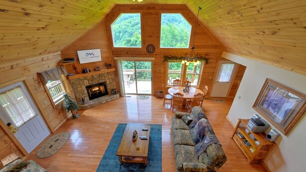 A spacious loft-style room with a stone fireplace, wooden floors, a large sofa, and a dining area with a view of the outdoors. - Pet-Friendly Modern Cabin w/ Hot Tub, Mountain Views + Biltmore Pass (Waynesville)