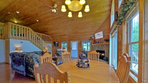 A wooden cabin interior with a dining table, chairs, a fireplace, and a staircase. - Pet-Friendly Modern Cabin w/ Hot Tub, Mountain Views + Biltmore Pass (Waynesville)