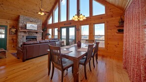 Un intérieur de chalet chaleureux avec une cheminée en pierre, une table à manger en bois et de grandes fenêtres donnant sur le paysage extérieur.