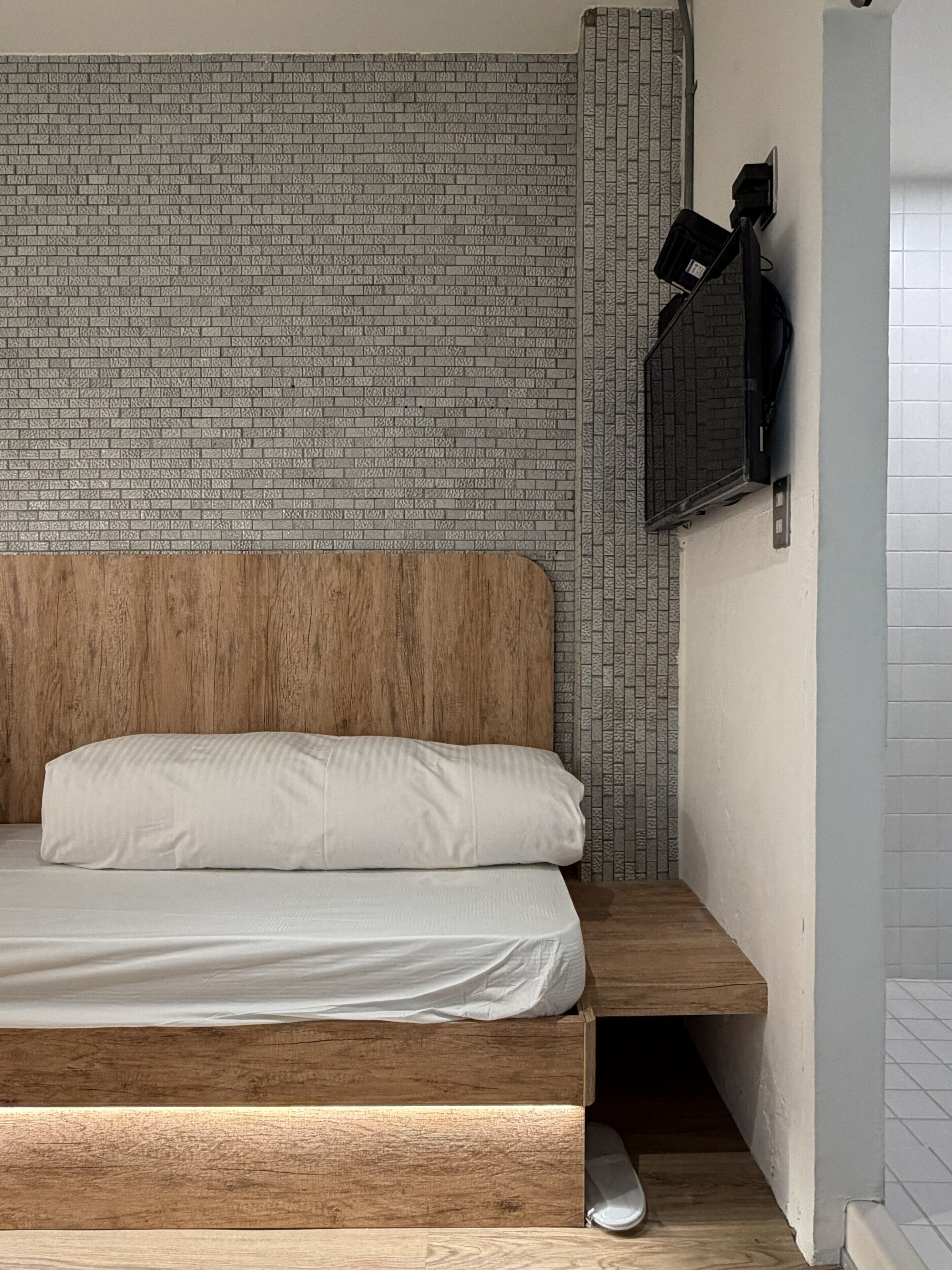 A bed with a wooden headboard and white bedding, a brick-patterned wall, and a mounted television.