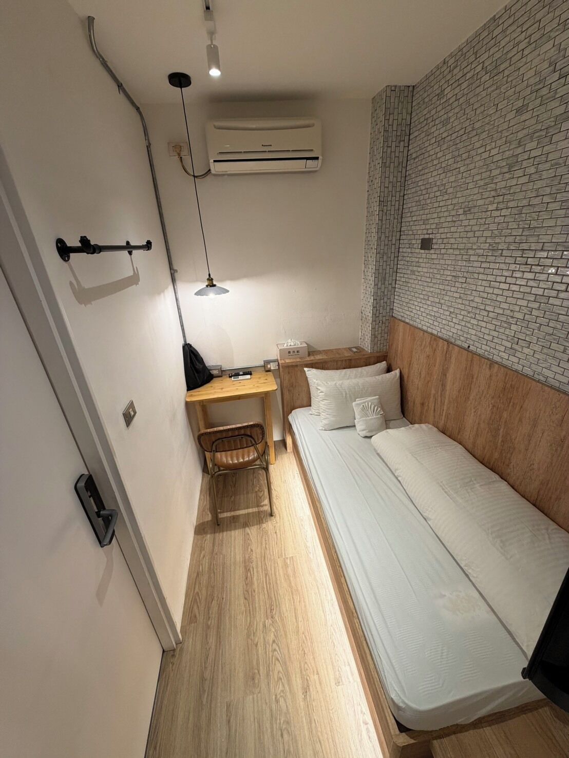 A compact room with a bed, a desk, a chair, an air conditioning unit, and a hanging lamp.