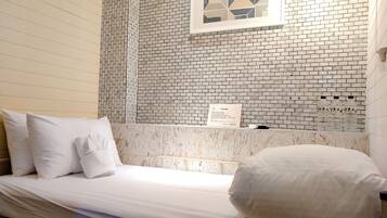 Quarto de hotel com cama, roupa de cama branca, parede decorada com mármore e uma obra de arte geométrica, além de um arranjo com garrafas de água.