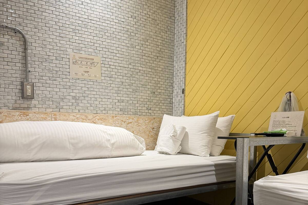 Quarto de hotel com parede em padrão de tijolo, parede de destaque amarela, cama com roupa de cama branca e uma pequena mesa com um objeto verde.