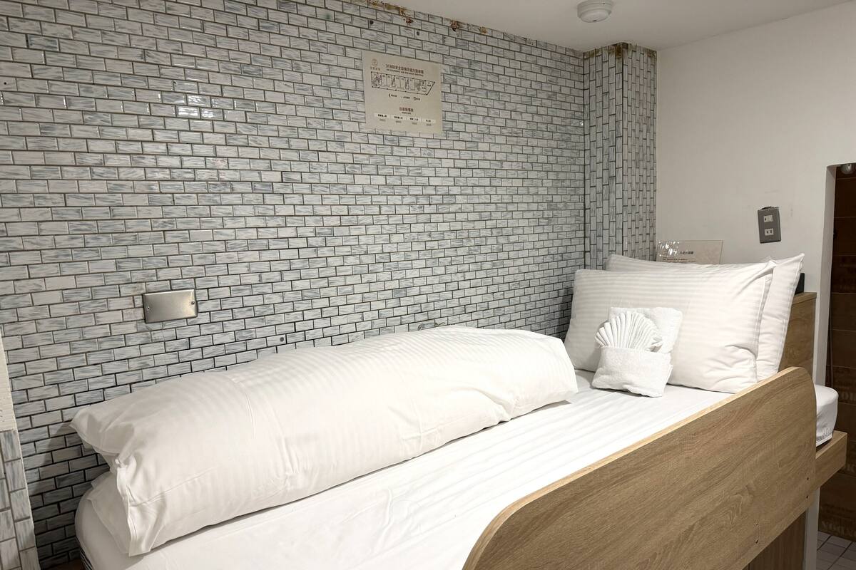 Uma cama de solteiro com cabeceira de madeira, roupa de cama branca e parede com padrão de tijolos.