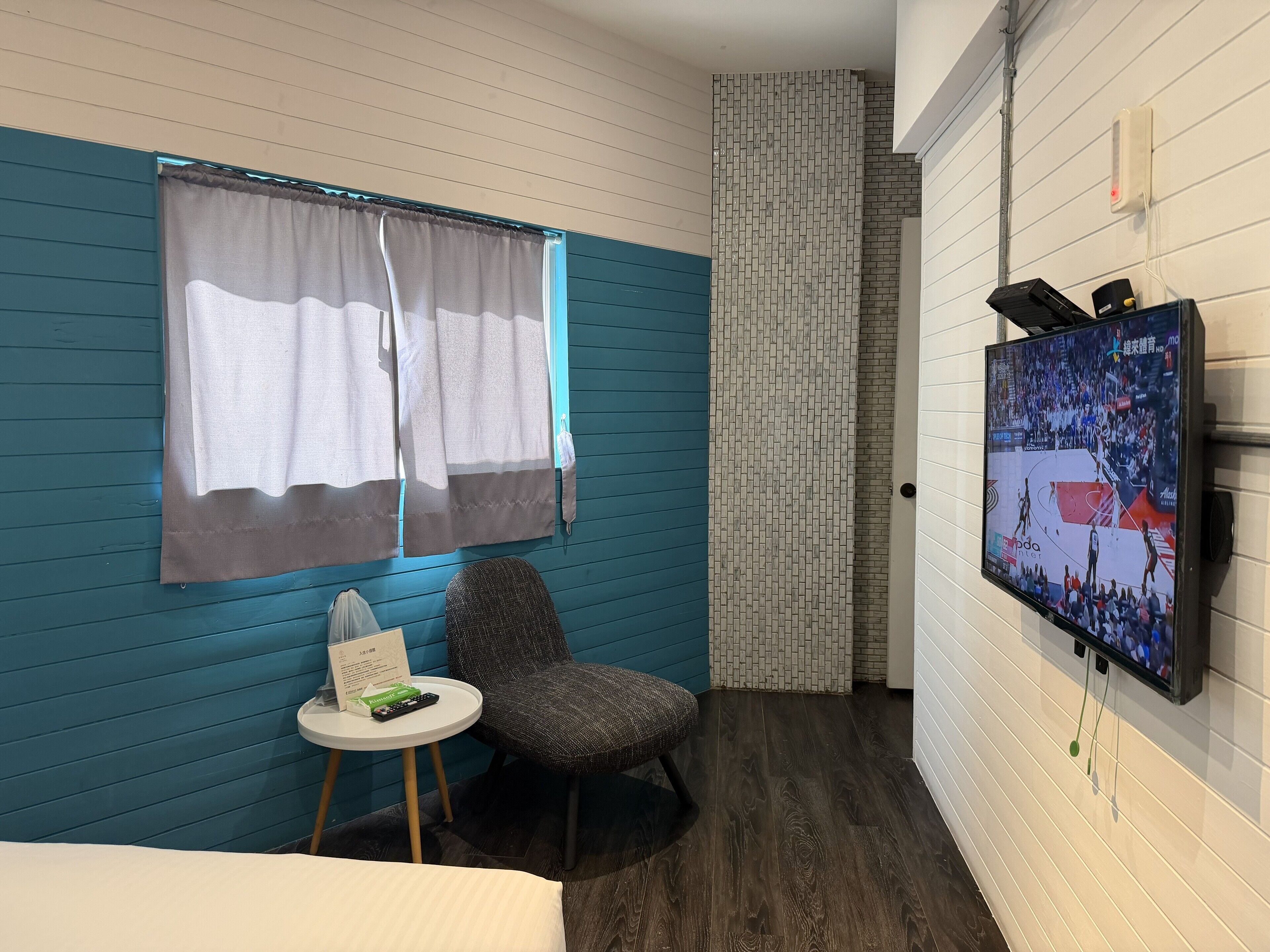 Une chambre d’hôtel aux murs de couleur turquoise, avec un lit, une petite table ronde sur laquelle se trouve une télécommande, une chaise et un téléviseur fixé au mur qui diffuse un événement sportif.