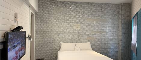 Habitación de hotel moderna con pared de ladrillo, televisor de pantalla plana montado en la pared, cama bien hecha y un pequeño cubo de basura.