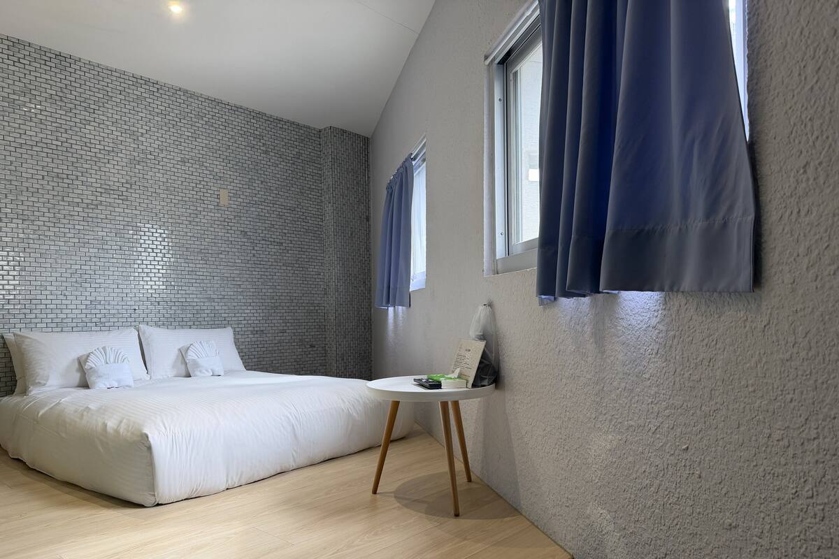 Quarto de hotel moderno com parede em padrão tijolo, uma cama grande, uma mesa redonda pequena e uma janela com cortinas.