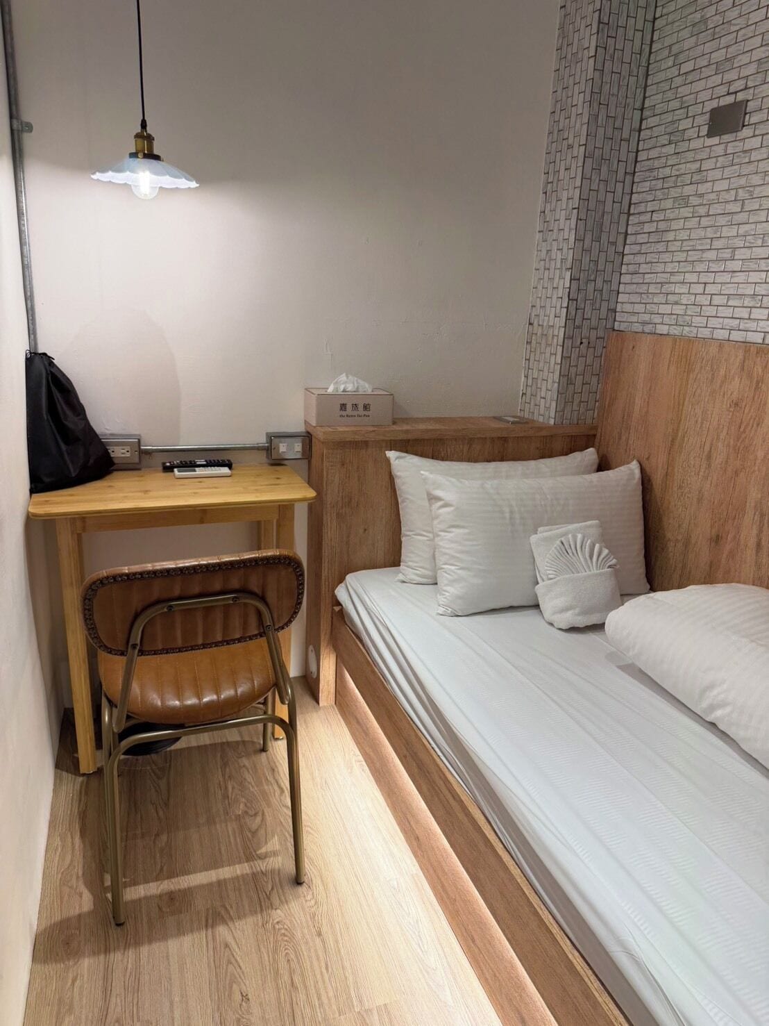 A compact hotel room with a wooden desk, a single bed, a chair, and a wall-mounted shelf.