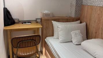 A compact hotel room with a wooden desk, a single bed, a chair, and a wall-mounted shelf.