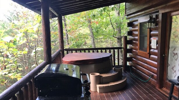 Una terrazza in legno con barbecue, vasca idromassaggio e zona salotto esterna, immersa tra gli alberi.