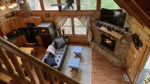 Interno di una baita con camera a soppalco, zona living con camino, cucina e ampie finestre che si affacciano su una foresta.