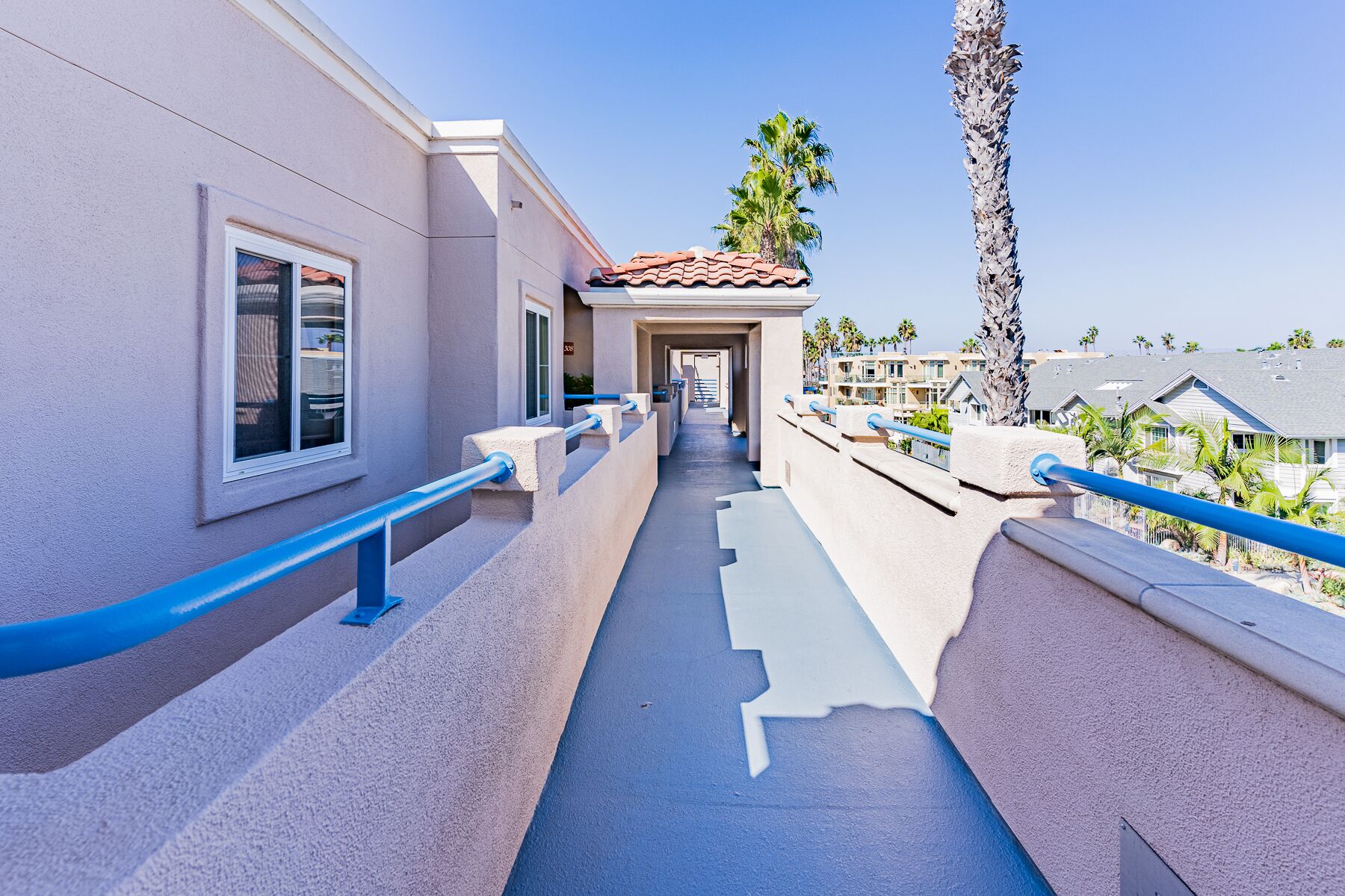 A corridor with a blue railing, a tiled roof, and a view of palm trees and houses in the background.
