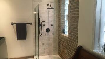 A modern bathroom with a glass-enclosed shower, a marble-tiled wall, a brick accent wall, and a window with a view.