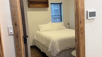 Quarto com porta de madeira, cama com roupa de cama branca, pequena janela e luminária fixada na parede.