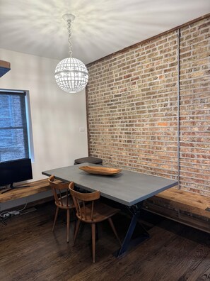 Een eetgedeelte met een bakstenen muur, een moderne kroonluchter en een houten tafel met stoelen.