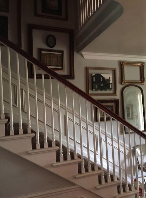 A staircase with white balusters and a wooden handrail, leading to a landing with framed artwork and a mirror.