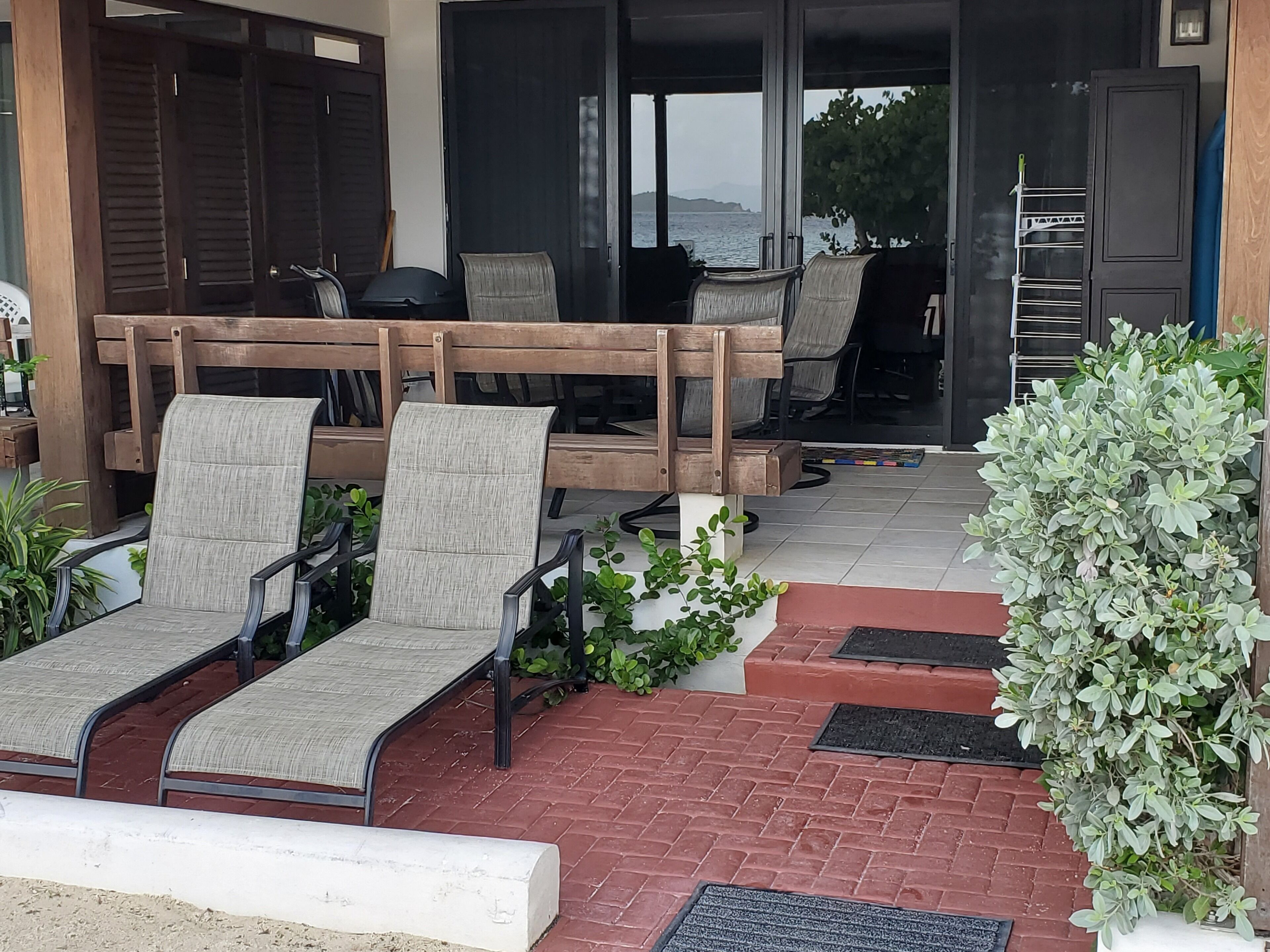 A covered outdoor seating area with cushioned chairs and a wooden bench, a potted plant, and a view of the sea through the open door.