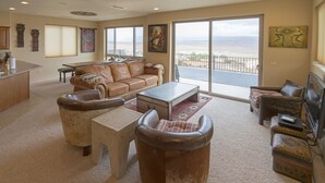 Amplia sala de estar con sofá de cuero, una mesa de centro y televisor. Un gran ventanal ofrece vistas a la playa.