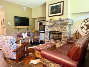 A living room with a stone fireplace, leather sofa, patterned armchair, and a television mounted on the wall.