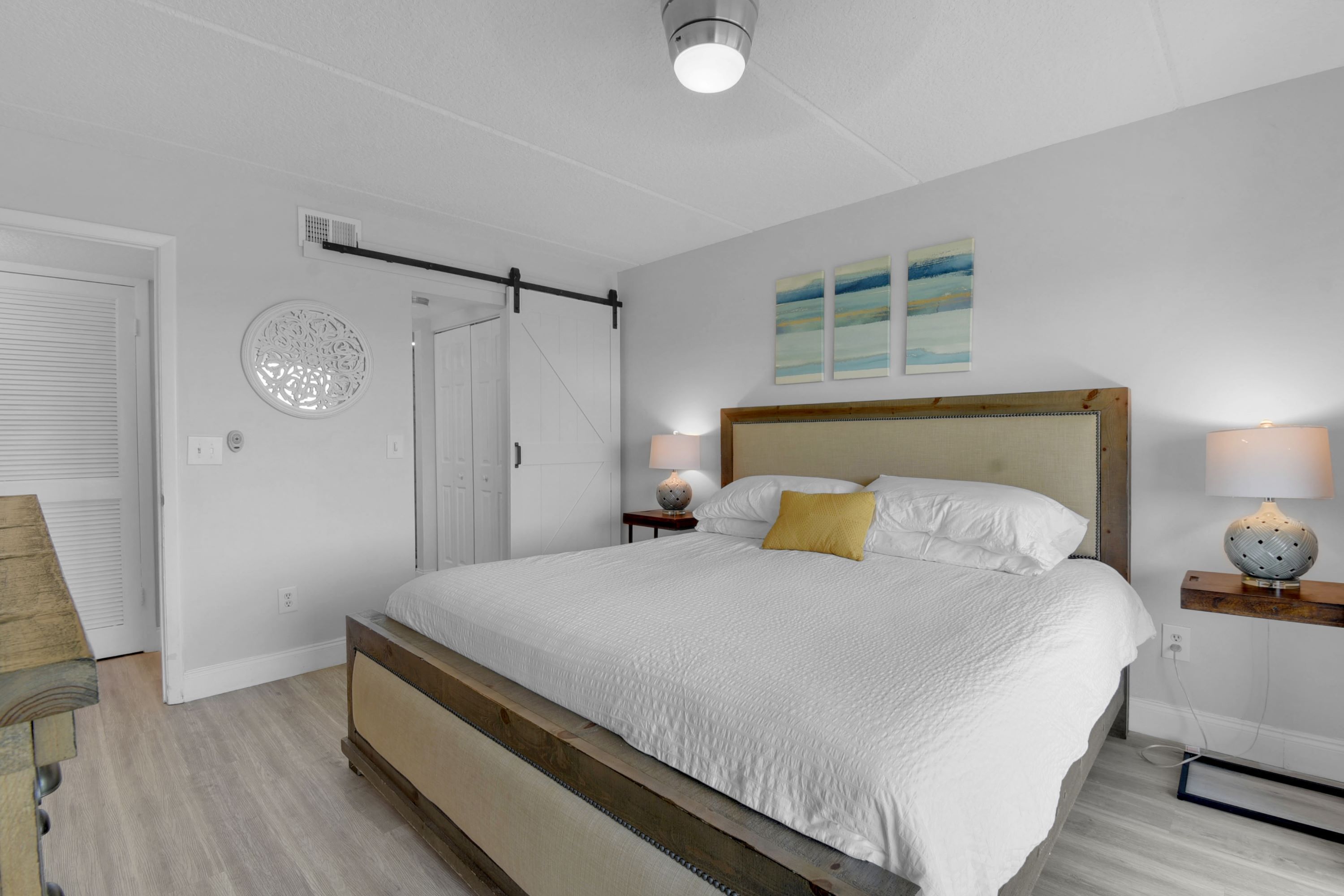 A bedroom with a large bed, a wooden headboard, a bedside table with a lamp, and a wall-mounted shelf.