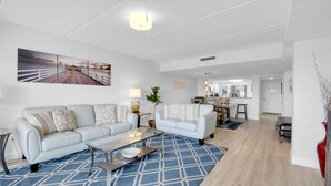 A modern living room with a sofa, coffee table, and a blue patterned rug. There is a dining area with a table and chairs in the background. - Ocean Front 2 Bed 2 Bath With Ocean Front Pool, walk to restaurants (St. Augustine)