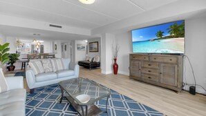 A modern living room with a large flat-screen TV displaying a beach scene, a glass coffee table, a sofa, and a dining area with a wooden table and chairs. - Ocean Front 2 Bed 2 Bath With Ocean Front Pool, walk to restaurants (St. Augustine)