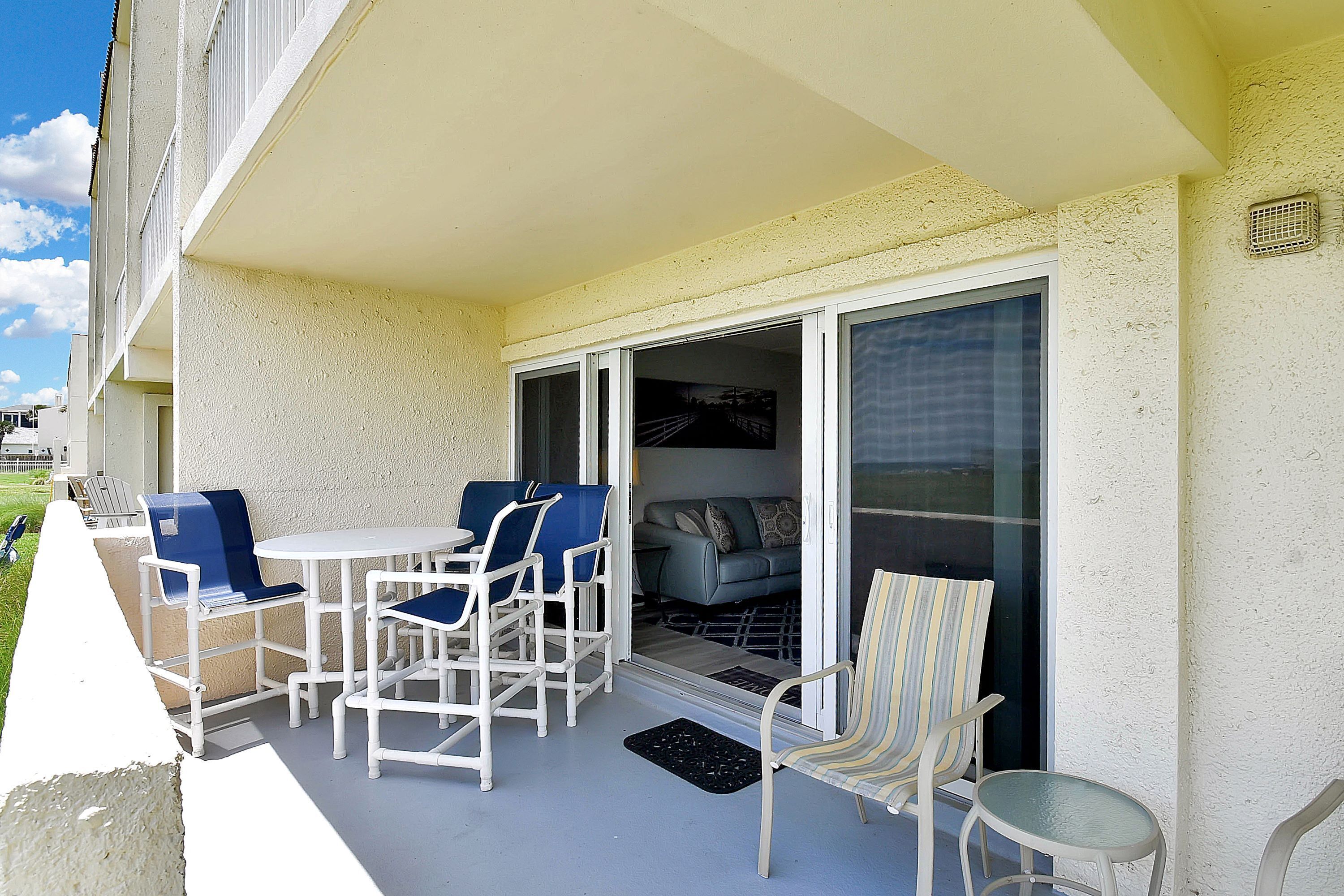 A balcony with white furniture, including a table and chairs, and a sofa inside a sliding glass door.