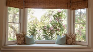 Un ventanal con asiento, dos almohadas y vistas a flores rosadas y follaje verde.